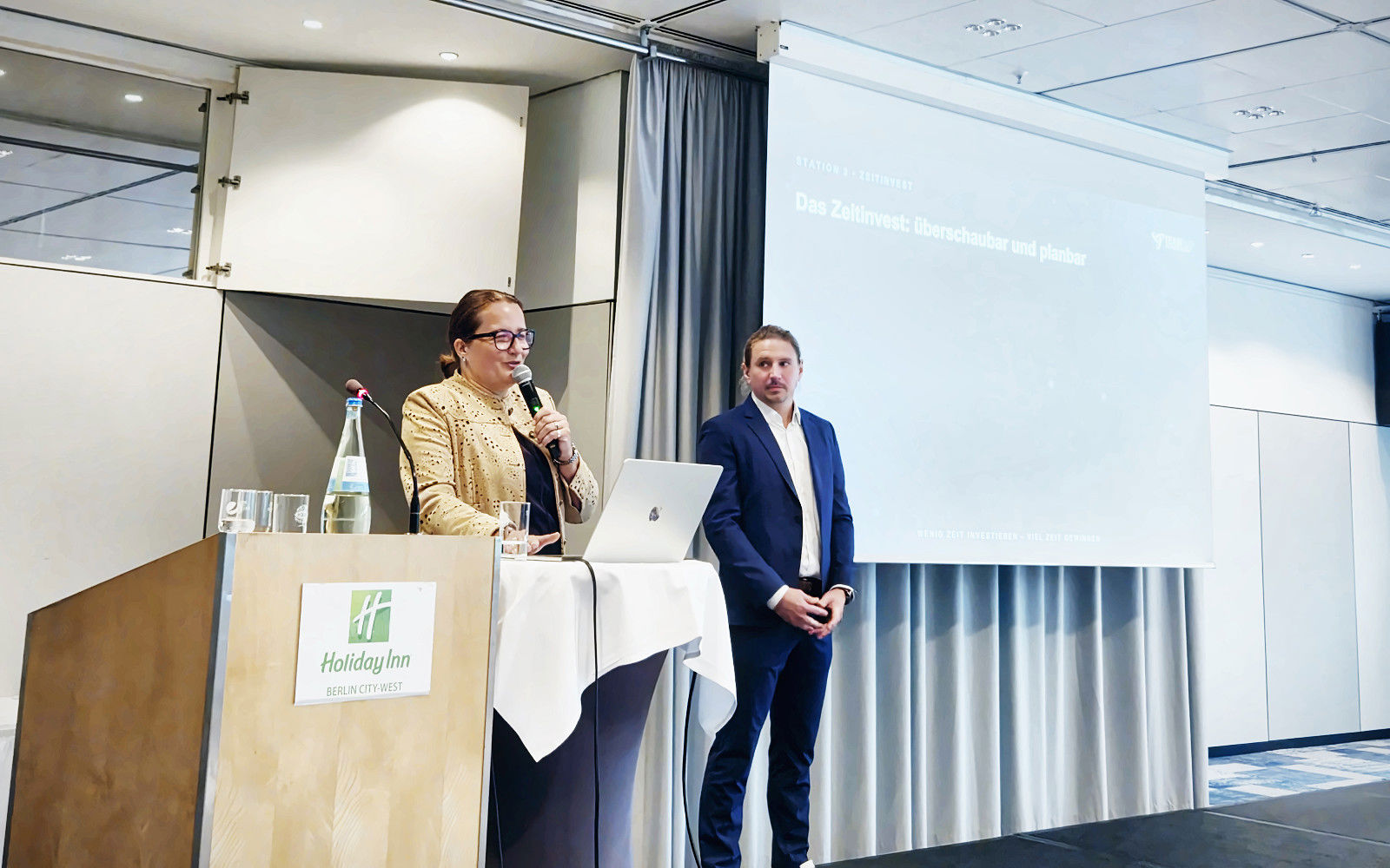 Alexandra Mollenkopf am Rednerpult und Max Fietkau auf der Bühne der Fachtagung in Berlin, im Hintergrund die Präsentation